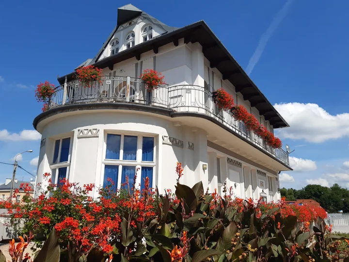 Na zdjęciu widać budynek z jasną elewacją, budynek z lat 20tych ubiegłego wieku. Wokół budynku i na balkonach widać kwitnące czerwone kwiaty.