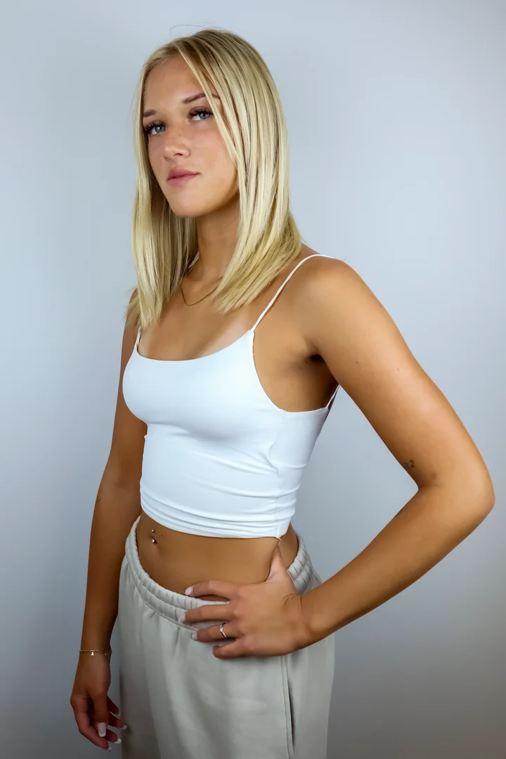 Portret młodej kobiety o blond włosach w jasnym topie, stojącej bokiem z ręką na biodrze, na neutralnym tle.