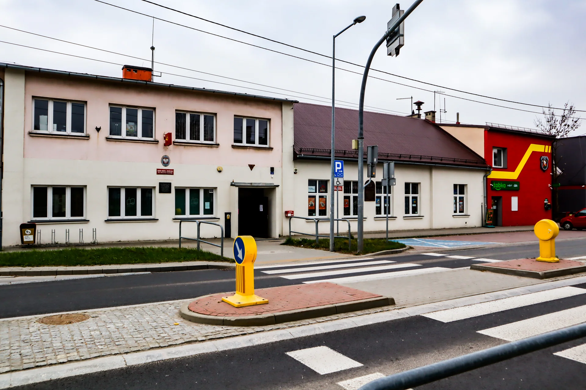 Budynek użyteczności publicznej przy ulicy z przejściem dla pieszych i sygnalizacją świetlną; obok widoczny czerwony fragment z wejściem do sklepu.