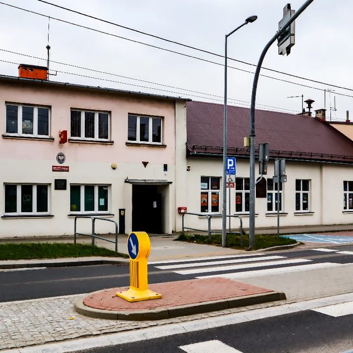Budynek użyteczności publicznej przy ulicy z przejściem dla pieszych, znakiem parkingu dla osób z niepełnosprawnościami i żółtą wysepką na środku jezdni.
