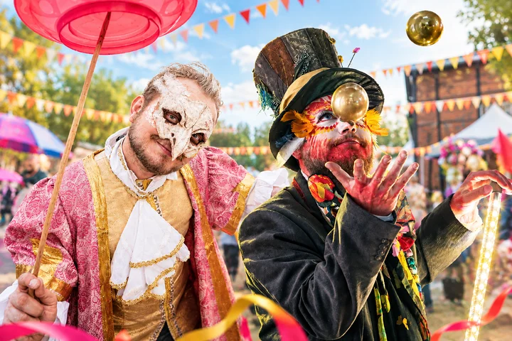 Artysta w masce kręcący talerzem oraz postać w cylindrze żonglująca złotymi kulami podczas kolorowego festiwalu plenerowego